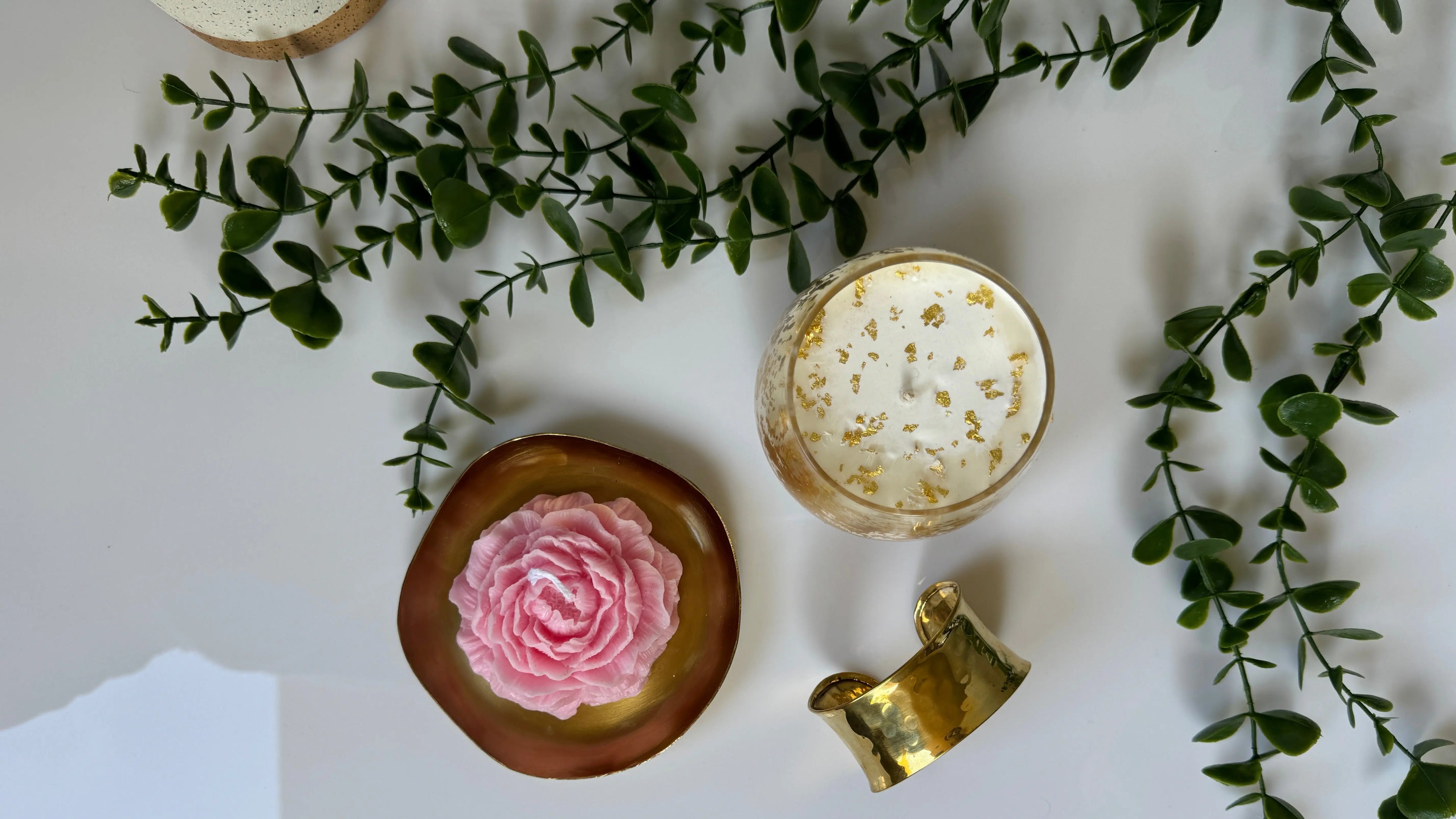 Shot from above, a pink peony shaped candle on a golden dish, a white candle with gold leaf flakes and a brass bangle